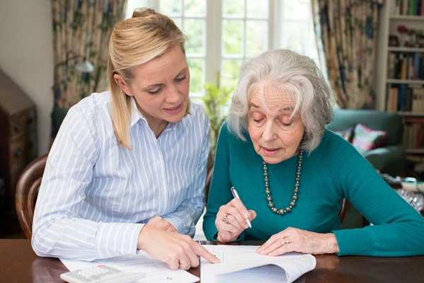 An elderly woman signing documents while a younger woman assists, pointing at the paperwork on a table in a well-lit home setting.